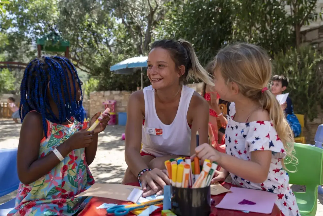 Une animatrice souriante encadre deux jeunes filles lors d'une activité créative en plein air.