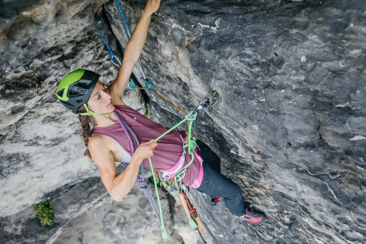 Donna con imbrago arrampicata uomo, casco verde, si arrampica su roccia grigia con corde colorate.