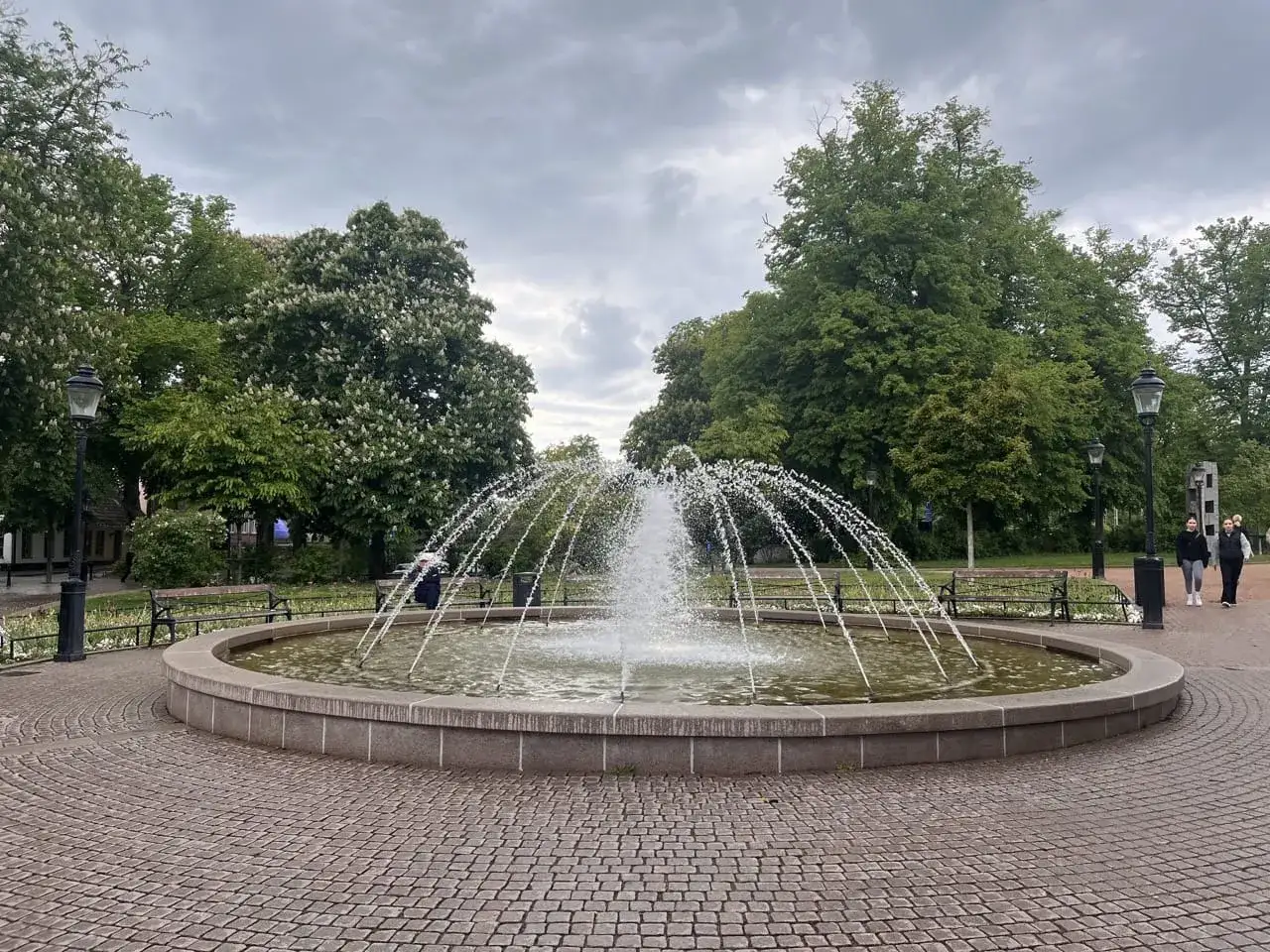 En fontän sprutar vatten i en park i Lund utomhus. Träd och bänkar omger fontänen.