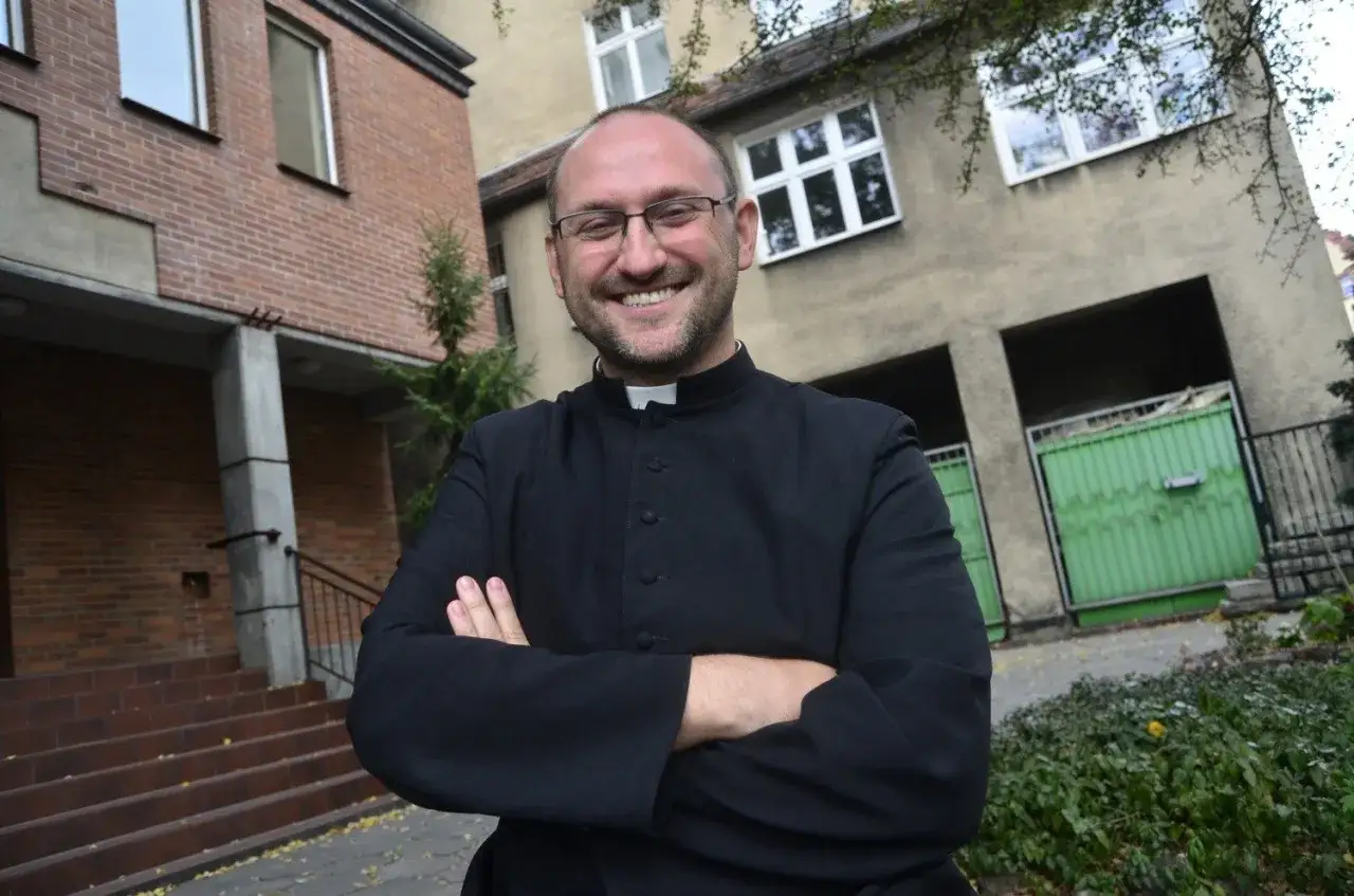 Uśmiechnięty ksiądz w sutannie, z rękami skrzyżowanymi na piersi, stoi przed budynkiem. Jego wybór celibatu jest świadectwem poświęcenia.