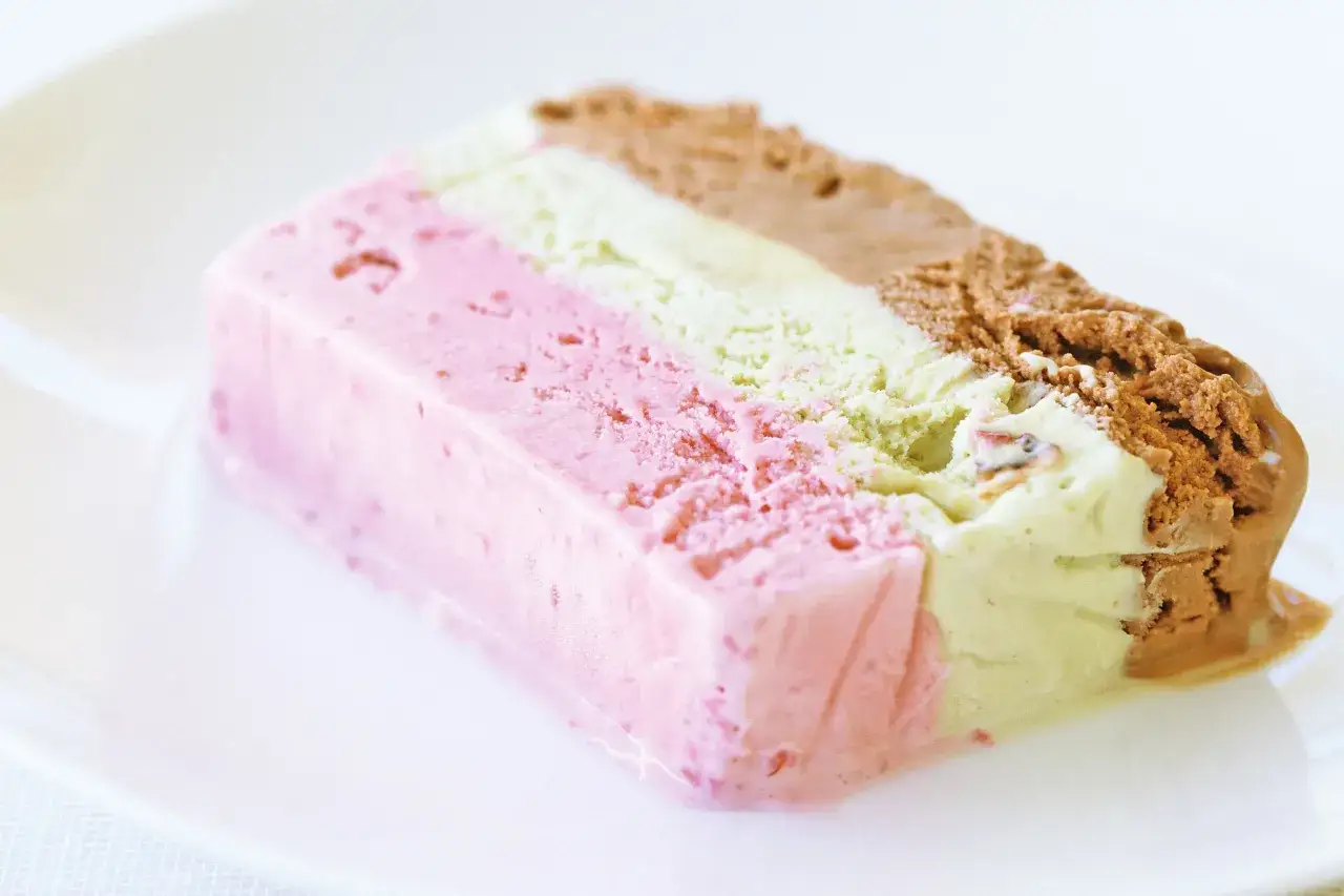 Trójwarstwowe lody cassate: różowe truskawkowe, pistacjowe i czekoladowe, na białym talerzu.