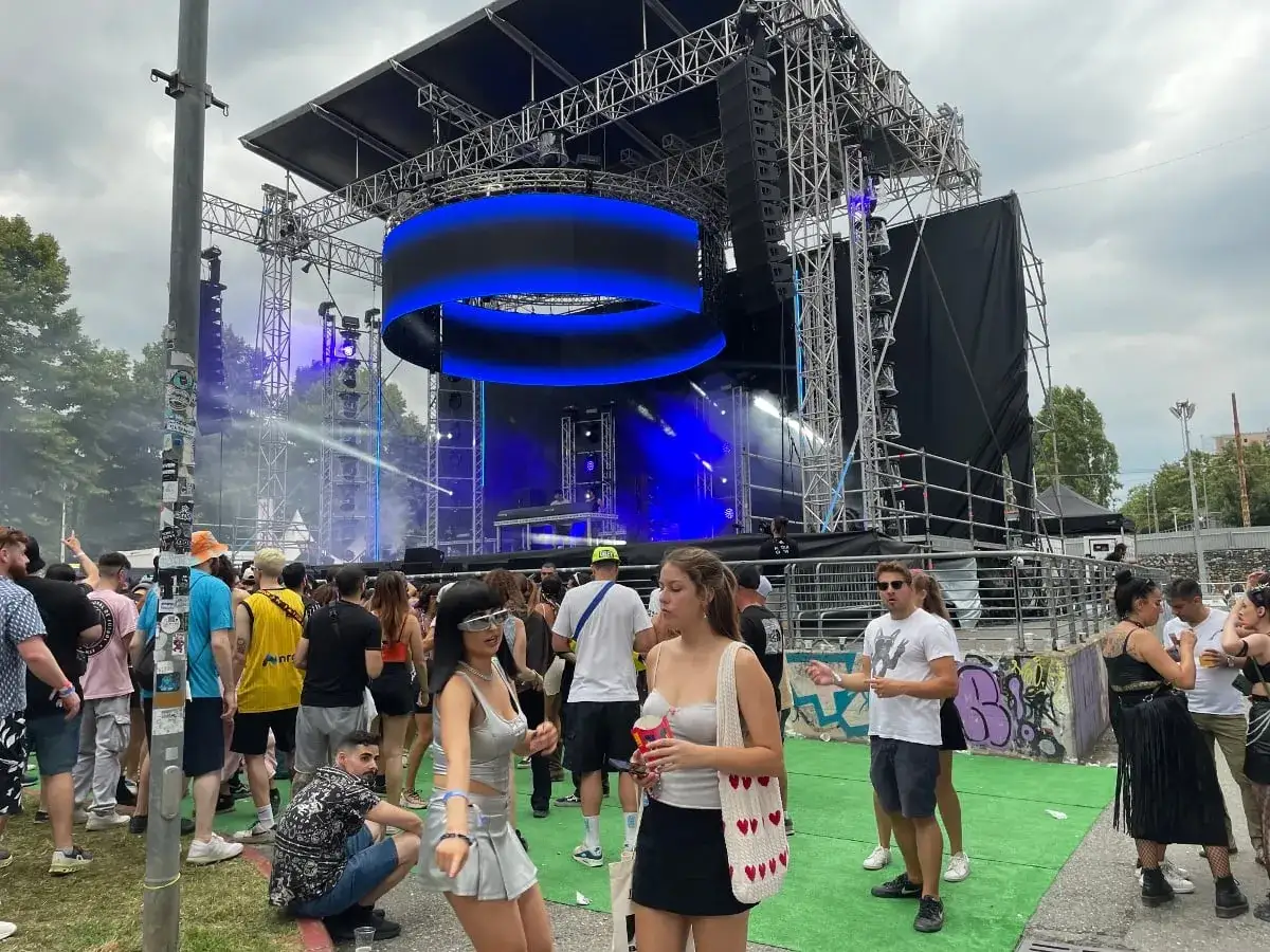 Folla in attesa di dj techno famosi sul palco illuminato da luci blu. Atmosfera da festival con gente che balla e si diverte.