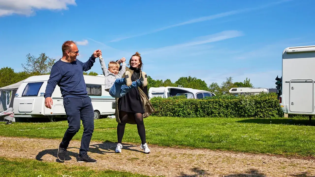 Familj njuter av en solig dag på camping i Billund. Pappa lyfter barnet medan mamman skrattar. Husvagnar och husbilar syns i bakgrunden.