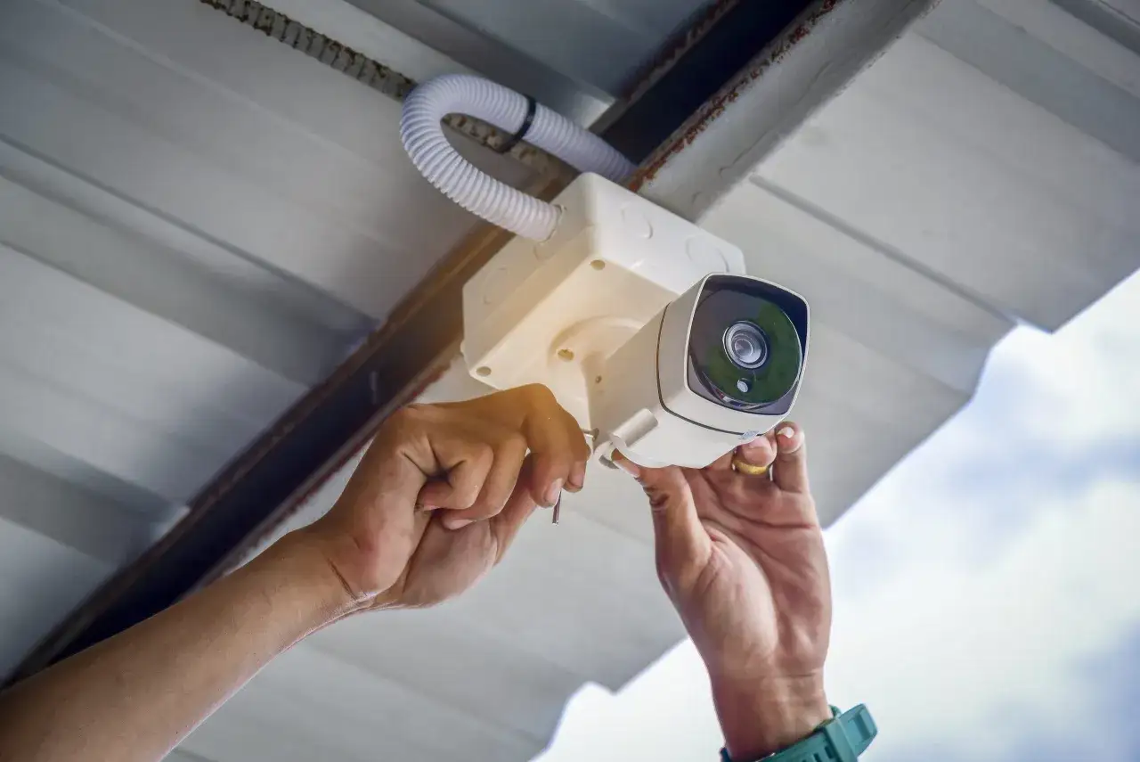 Installazione di un impianto di videosorveglianza: mani che fissano una telecamera a soffitto, con cavi e tubo protettivo visibili.