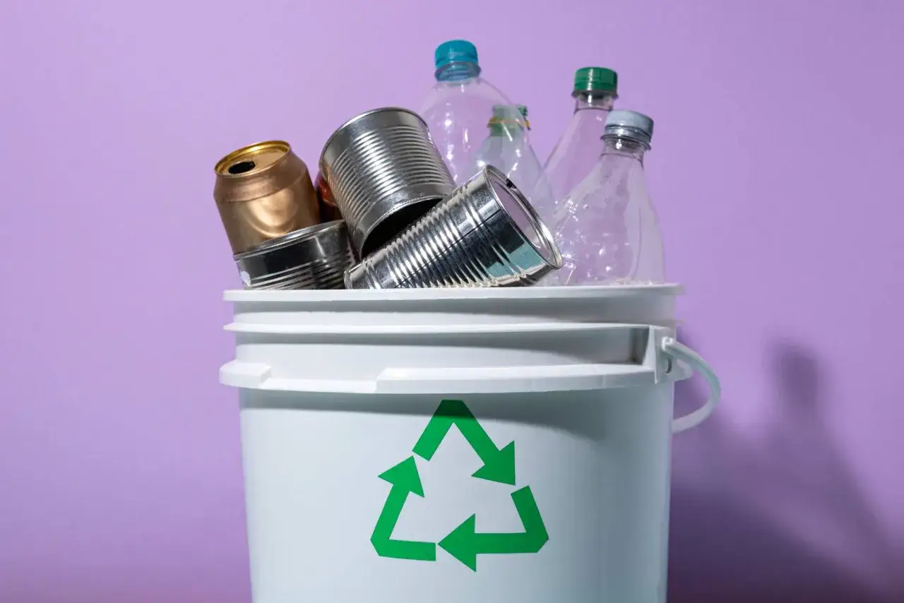 Biały pojemnik z zielonym symbolem recyklingu, wypełniony puszkami i plastikowymi butelkami. To pokazuje, co to jest recykling.