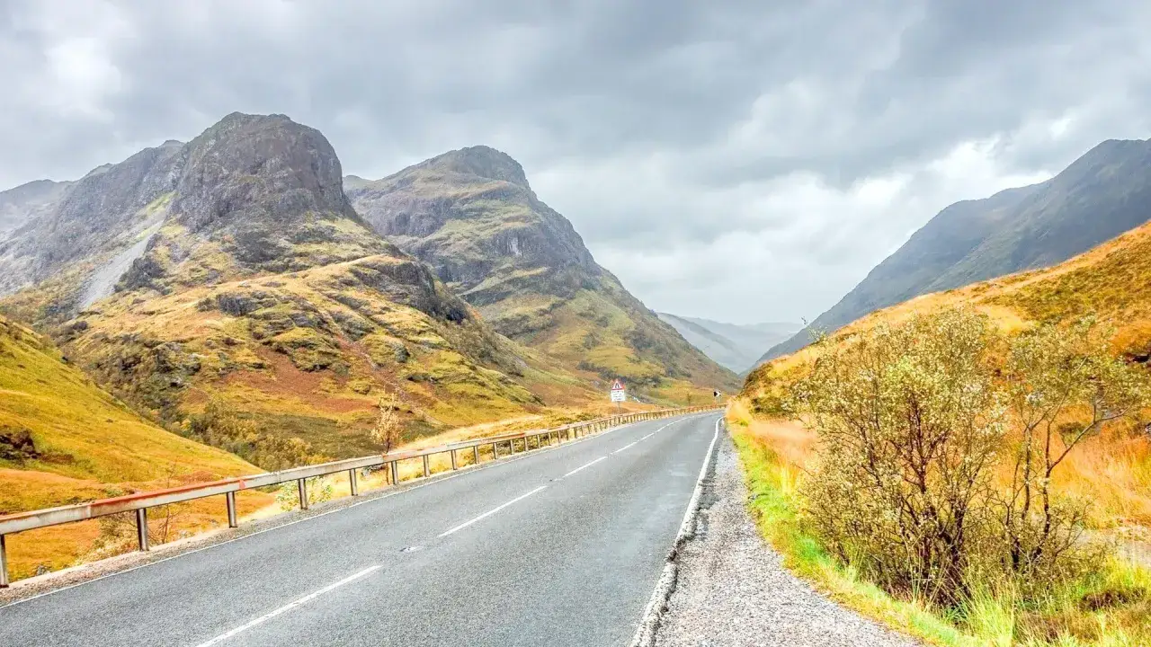 A winding road through a dramatic Scottish glen, perfect for a bike tour Scotland adventure. Mountains rise on either side, covered in autumnal hues under a cloudy sky.