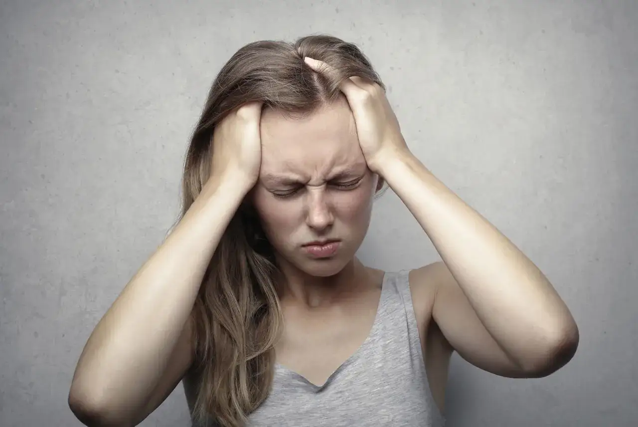 Una giovane donna con le mani sulla testa, gli occhi chiusi e un'espressione di dolore. Sembra tormentata da pensieri paranoici e pericolosi.