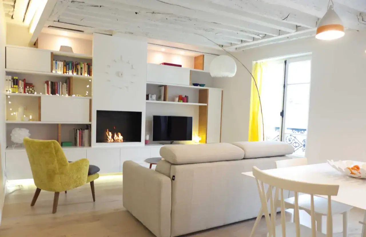 Salon lumineux d'un f3 appartement, avec cheminée, canapé, fauteuil jaune et étagères remplies de livres.