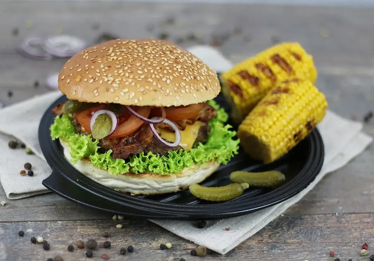 Pyszne burgery z grilla z chrupiącą sałatą, pomidorem i ogórkiem. Obok grillowana kukurydza i pikle.