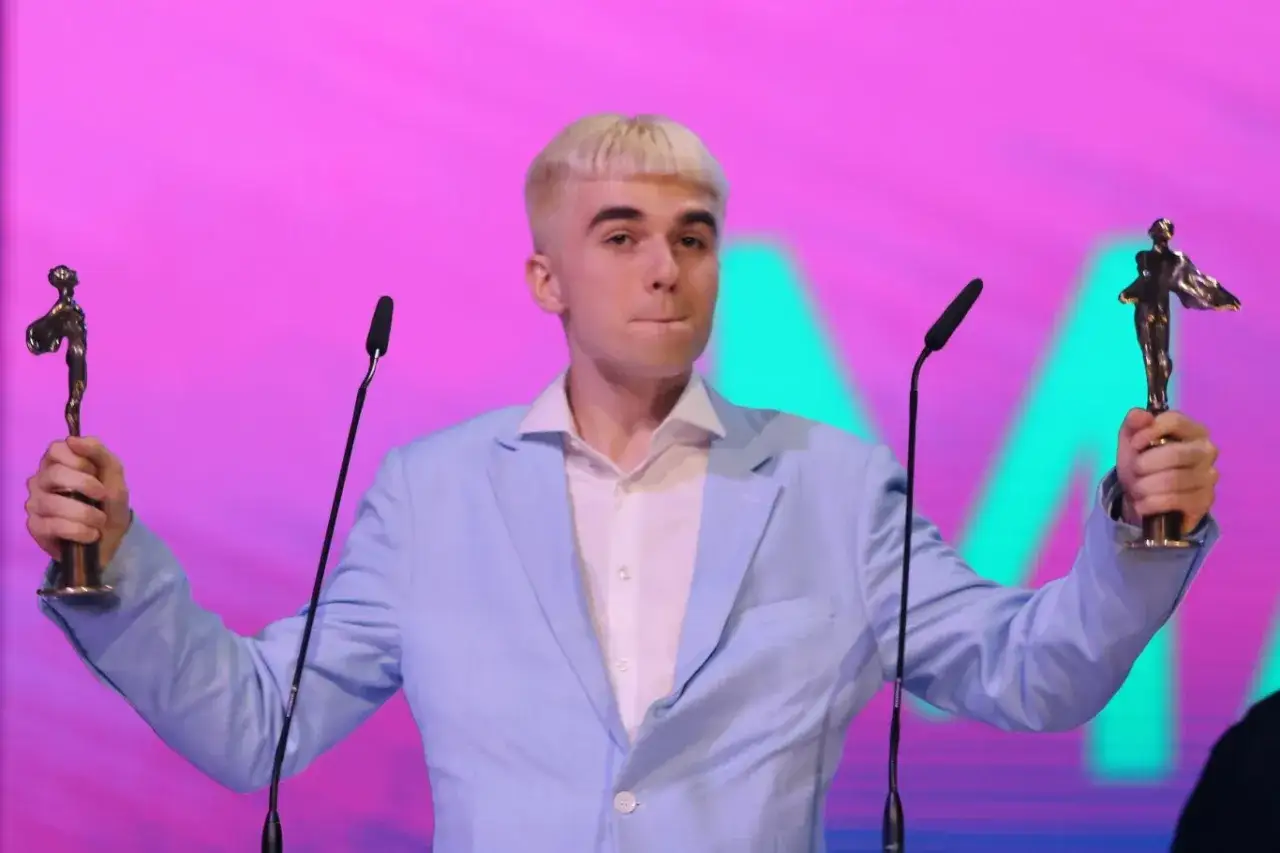Mc Mata z dwoma statuetkami na scenie, w jasnoniebieskiej marynarce.