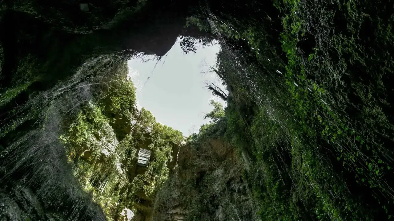 Vista desde abajo del **pozo de los aines**, con paredes cubiertas de vegetación exuberante y cascadas de agua cayendo hacia el cielo abierto.
