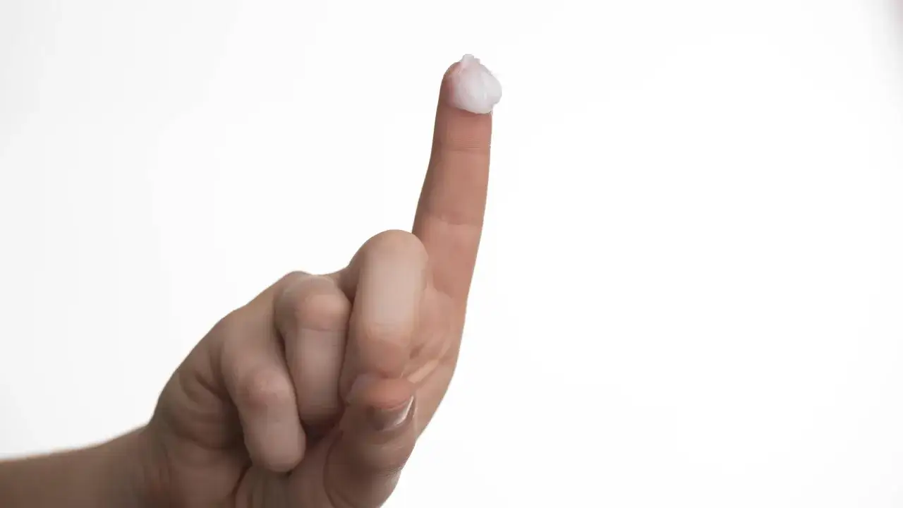 Ein Finger h&auml;lt eine kleine Menge wei&szlig;er Salbe, die f&uuml;r die Anwendung im Intimbereich gedacht ist.