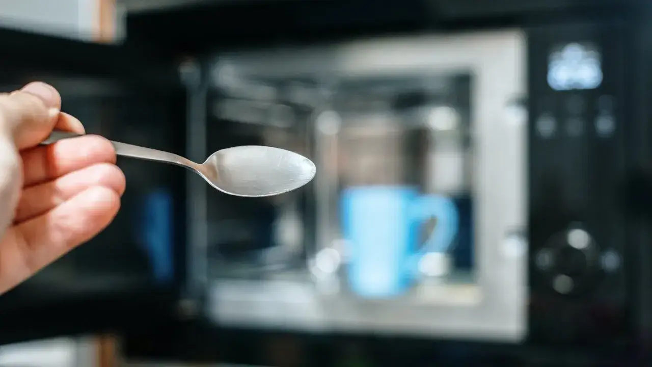 Hand hält Löffel aus Metall vor Mikrowelle mit blauer Tasse. Achtung: Metall in Mikrowelle ist gefährlich!