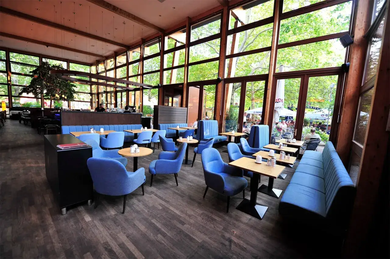 Gem&uuml;tliches **Cafe Wilmersdorf** mit blauen Sesseln und B&auml;nken, Holzboden und gro&szlig;en Fenstern, die auf eine gr&uuml;ne Terrasse blicken.