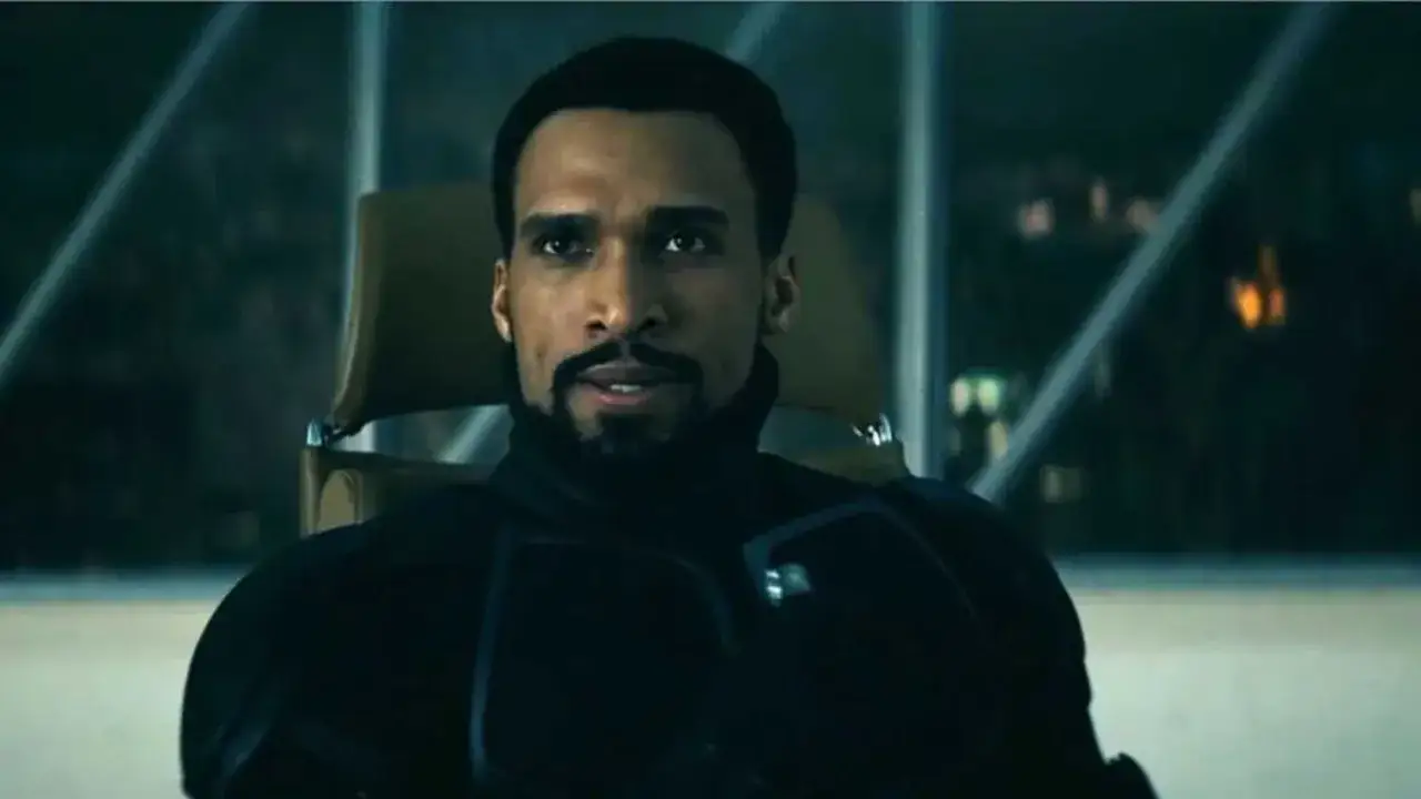 Un homme au regard intense, vêtu d'un costume noir, le **black noir sans masque**, est assis dans un fauteuil.