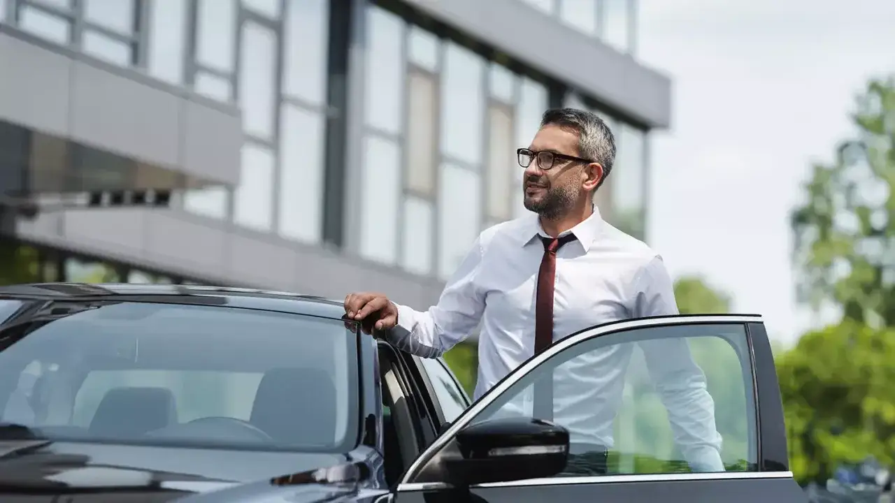 Biznesmen w okularach opiera się o lśniący, czarny samochód. Cieszy się z ulgi na samochód, która ułatwia mu życie.