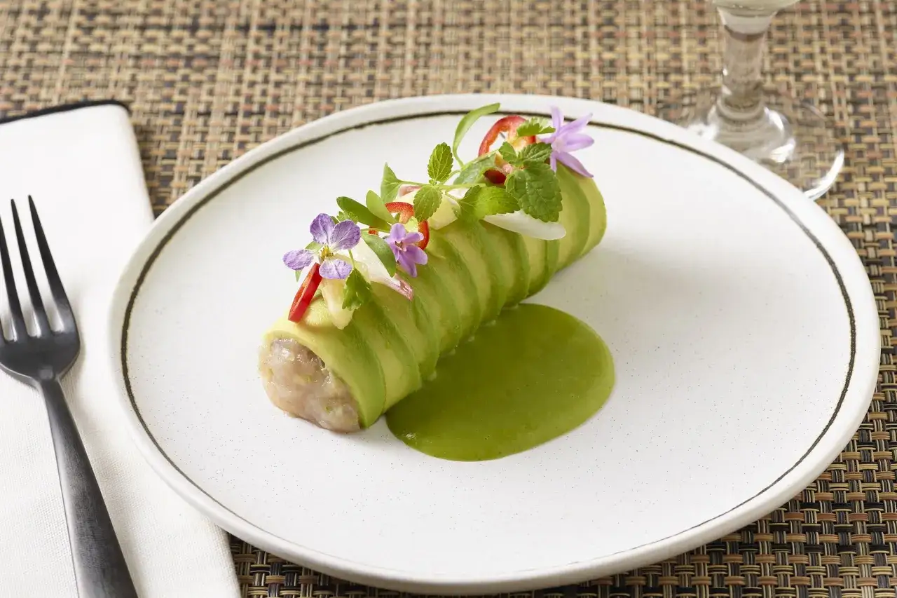 Un rouleau de tartare de thon frais, envelopp&eacute; d'avocat tranch&eacute;, garni de fleurs comestibles et servi avec une sauce verte vibrante.