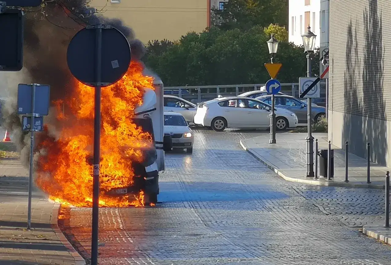Płonący samoch&oacute;d na ulicy, widać kłęby dymu i płomienie. Czy olej napędowy się pali? Inne auta stoją w pobliżu.