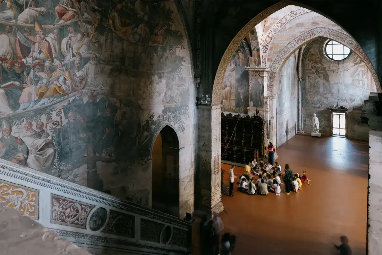 Interno dell'abbazia di Chiaravalle con affreschi storici e un gruppo di bambini seduti a terra, ascoltando una guida.