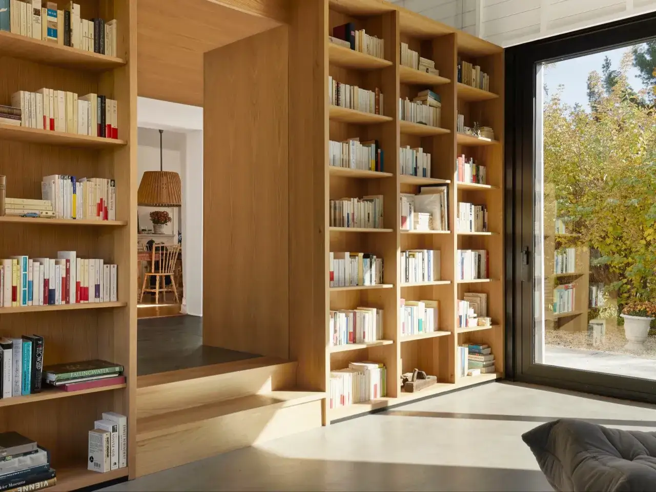 Przytulny gabinet biblioteka w domu, pełen książek na drewnianych półkach. Przez okno widać jesienny ogród.