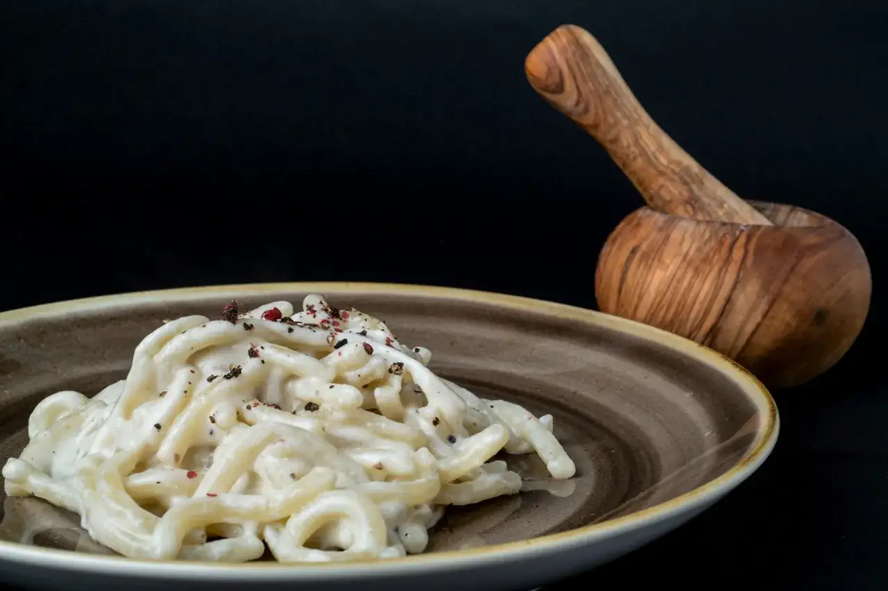 Un piatto di pasta cremosa, condita con pepe nero e rosa, evoca il piacere del mangiare toscano. Un mortaio in legno sullo sfondo.