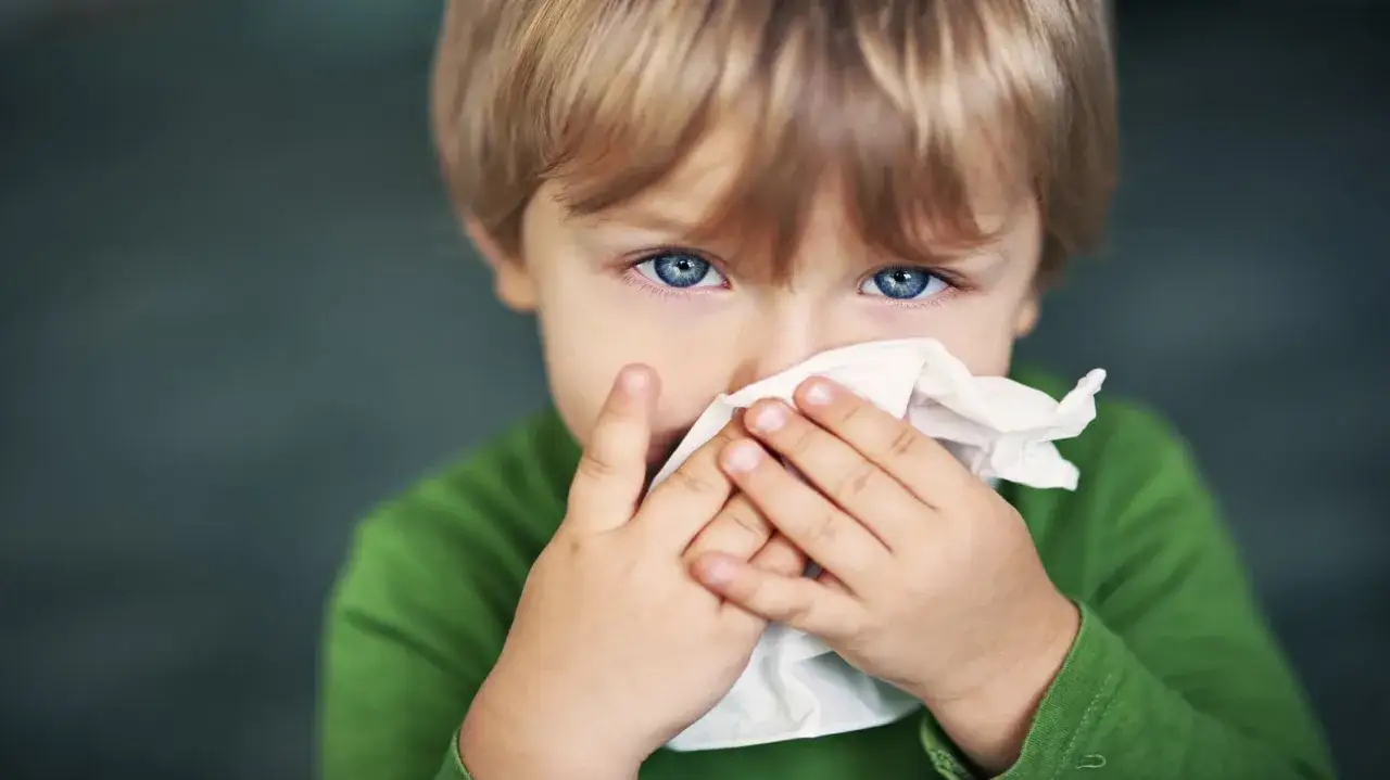 Bambino con occhi azzurri che si soffia il naso con un fazzoletto, forse per un po' di catarro in gola.