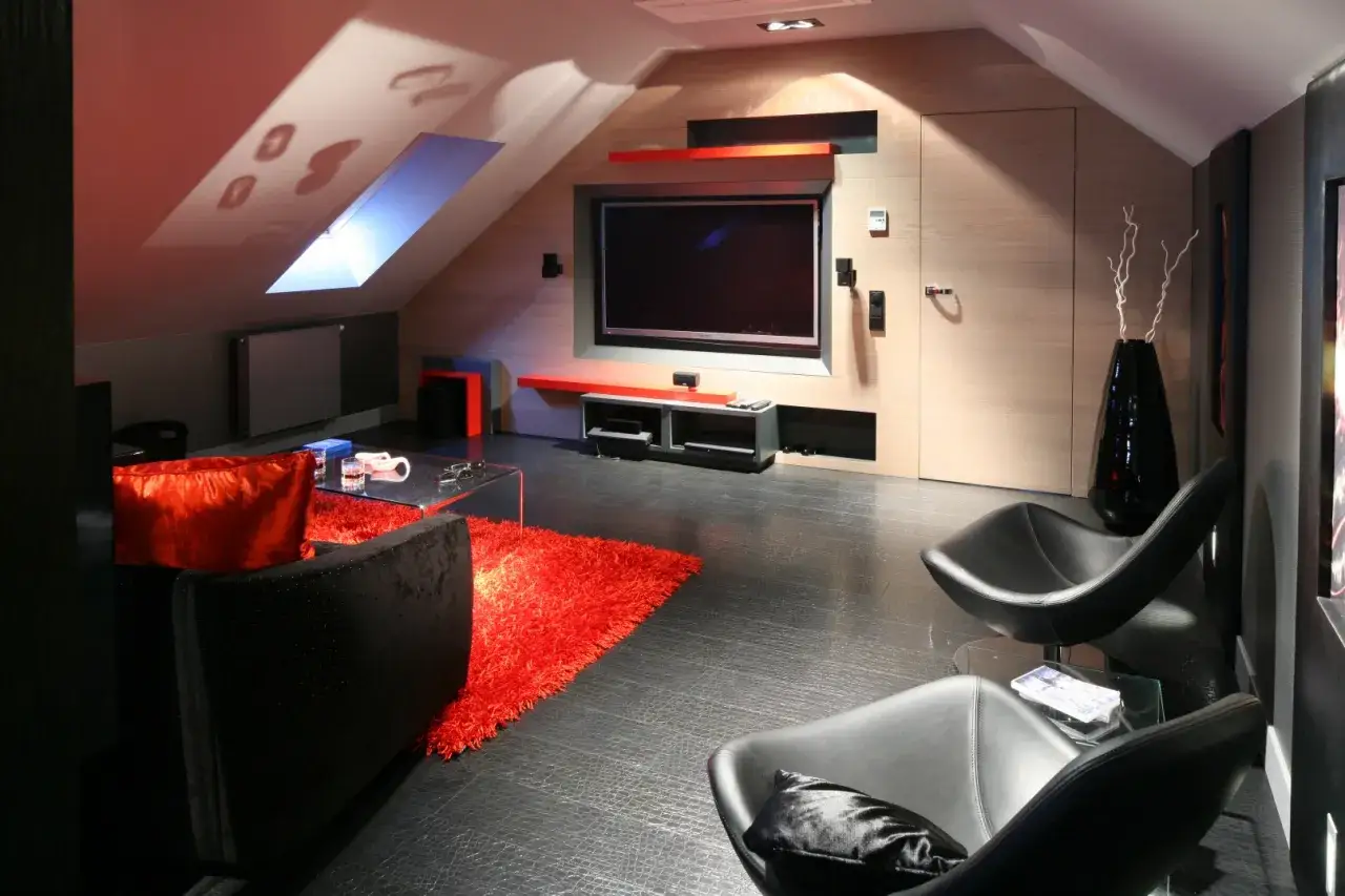 Nowoczesny salon na poddaszu ze skosami. Ciemna sofa, czerwony dywan i fotele tworzą przytulną przestrzeń do oglądania telewizji.