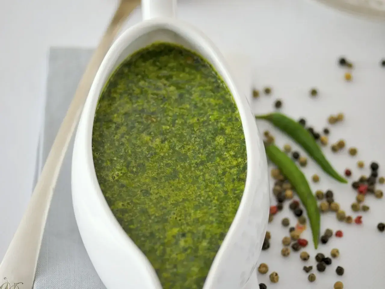 Gr&uuml;ne So&szlig;e in einer Sauciere, umgeben von Pfefferk&ouml;rnern und Chilis. Perfekt f&uuml;r Petersilie Rezepte.