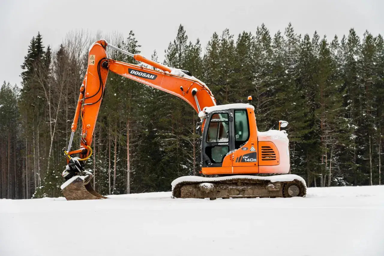 Pomarańczowa koparka Doosan DX 140R stoi na śniegu. To idealny sprzęt do pracy, nawet gdy pojawiają się pytania na egzamin koparki kl 1.