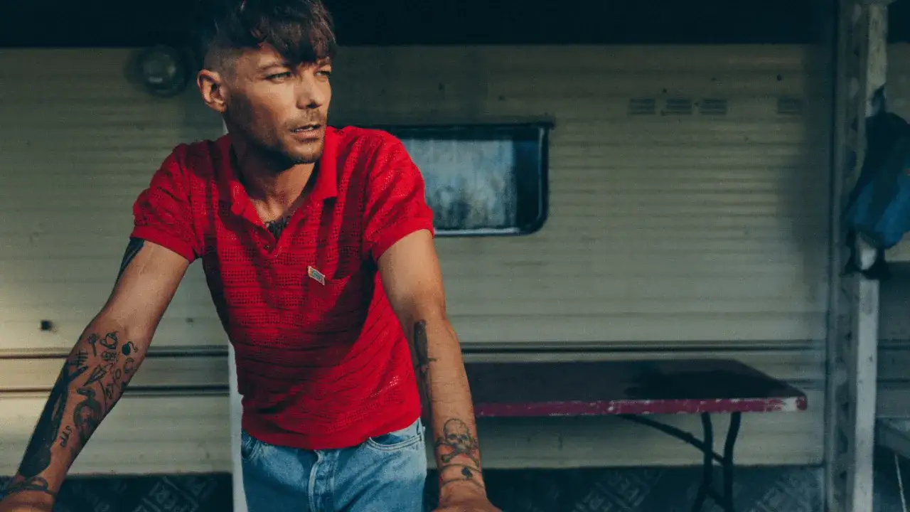 Louis Tomlinson w czerwonej koszulce polo i dżinsach, z tatuażami na ramionach, opiera się o przyczepę kempingową.
