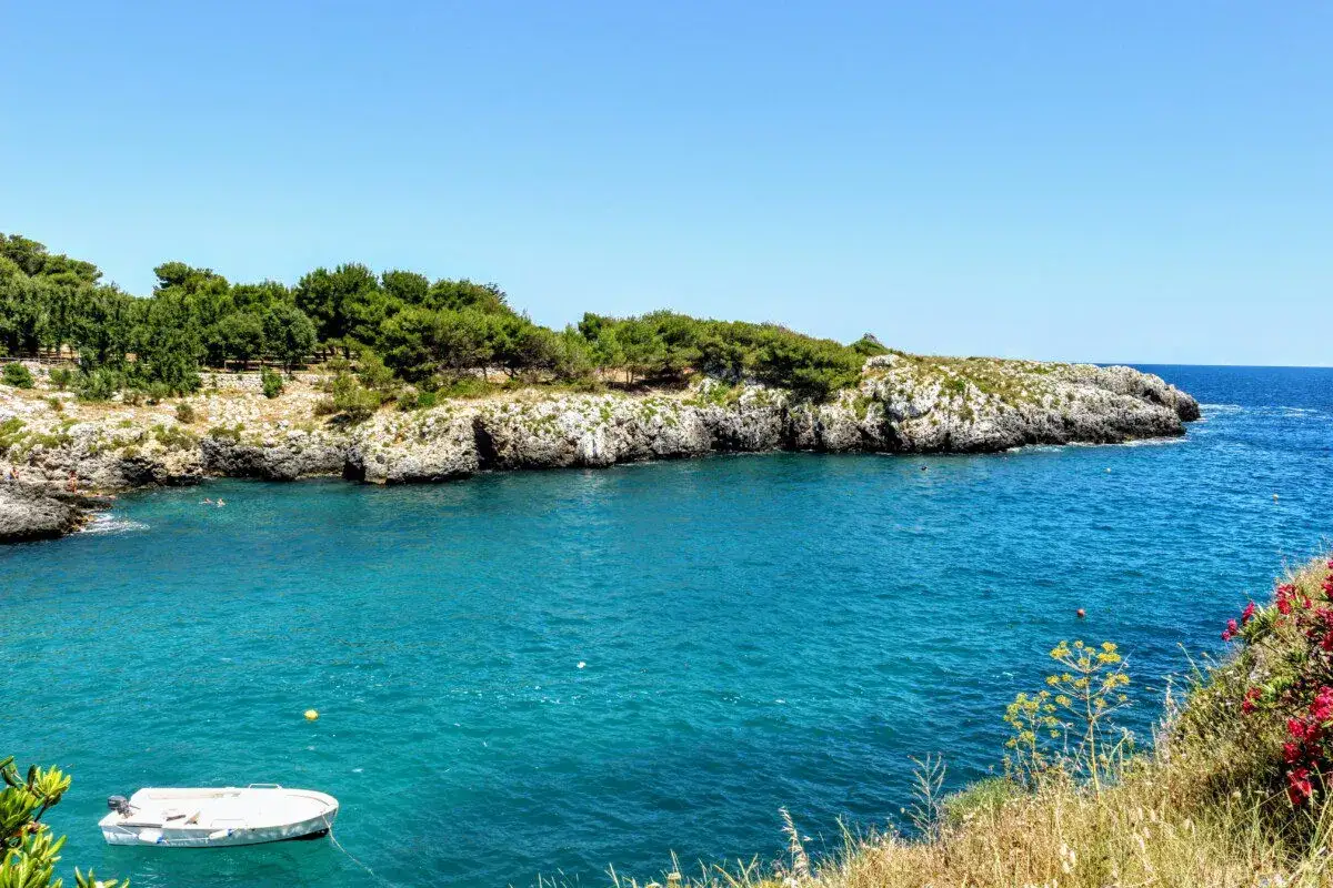 Acque cristalline e scogliere rocciose nel **Salento Ionico**. Una piccola barca bianca galleggia tranquilla, mentre il sole illumina la costa verdeggiante.