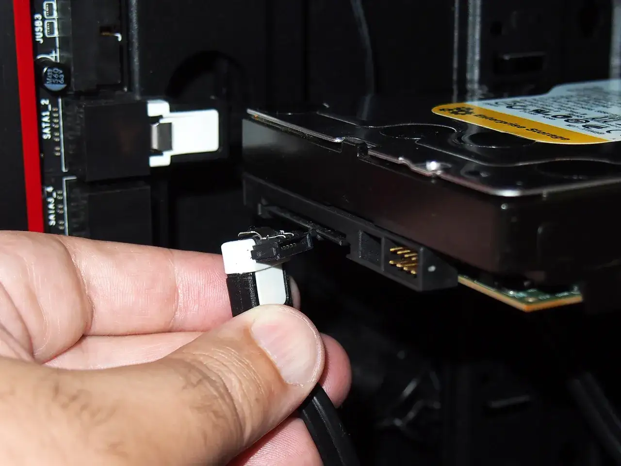 Connexion d'un câble à un disque dur serial ata. La main insère le connecteur blanc et noir dans le port du disque.