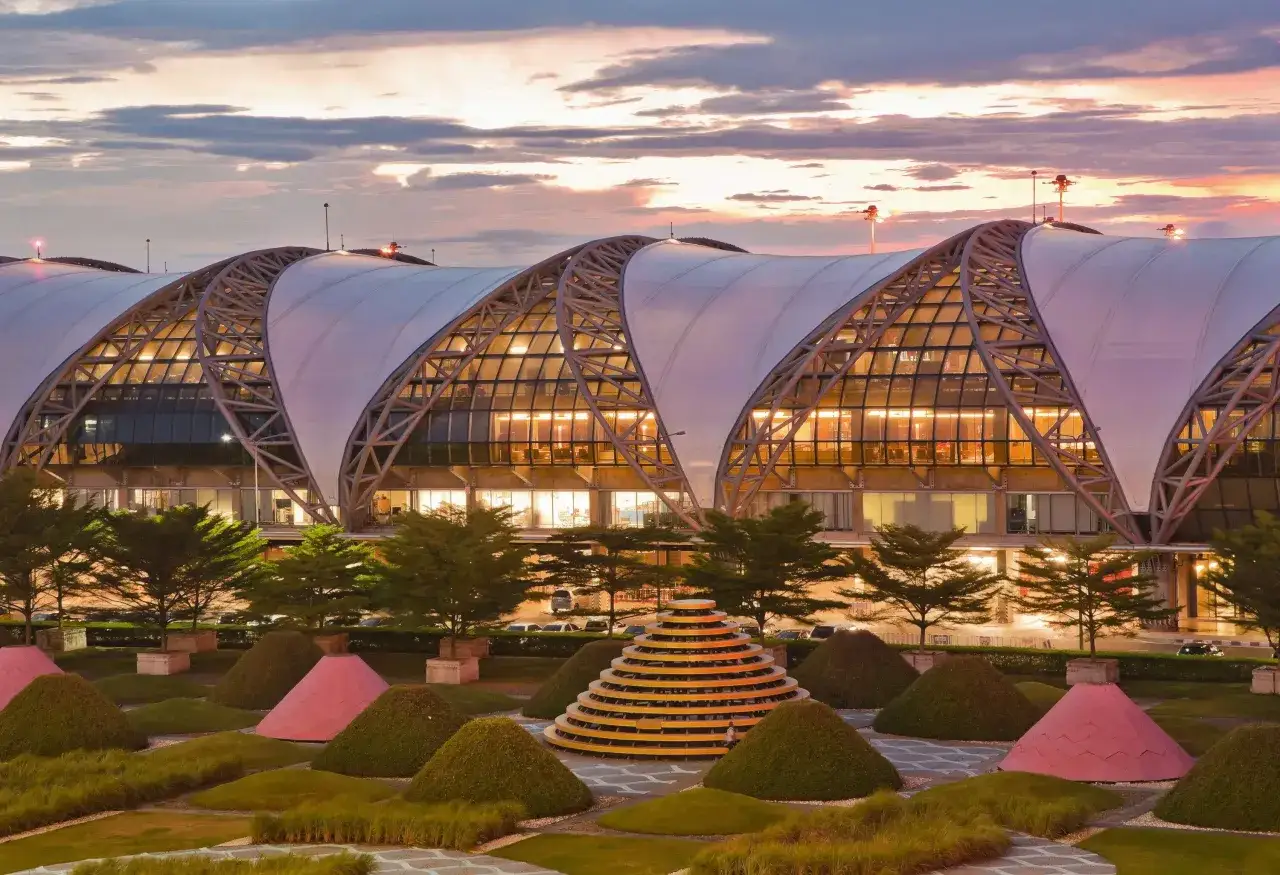 Der Flughafen Bangkok bei Sonnenuntergang. Die geschwungene Dachkonstruktion des Flughafens ist beleuchtet, davor ein Garten mit bunten Skulpturen und B&auml;umen.