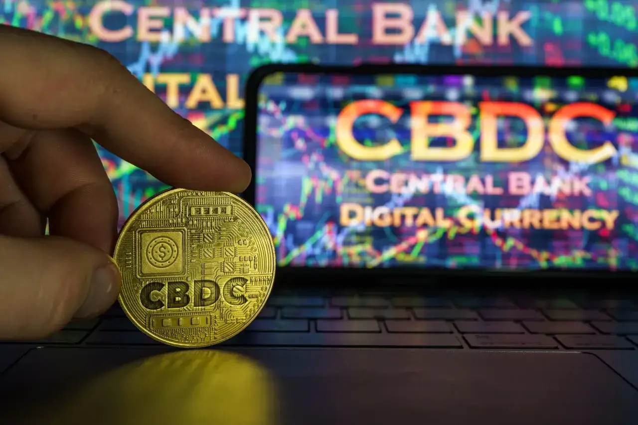 Złota moneta z napisem CBDC w dłoni, na tle ekranu z wykresem i napisem 