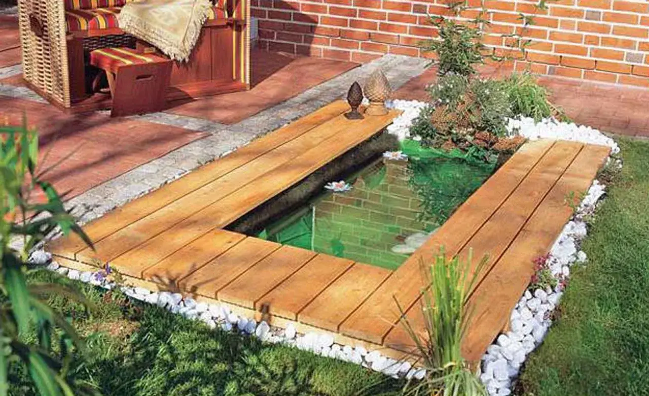 Ein moderner miniteich anlegen mit Holzdeck und Strandkorb. Weiße Kieselsteine umranden das grüne Wasser.