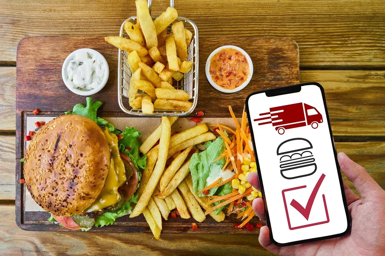 Hamburguesa, papas fritas y ensalada listos para llevar. El tel&eacute;fono muestra un cami&oacute;n de reparto y una hamburguesa, indicando que es un pedido para llevar.
