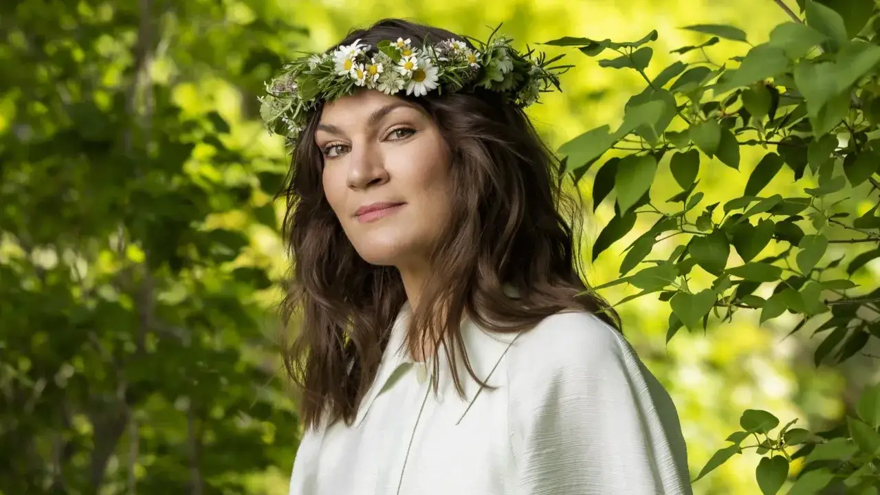 Malin Wollin bär en blomsterkrans i sitt bruna hår, omgiven av grönska.