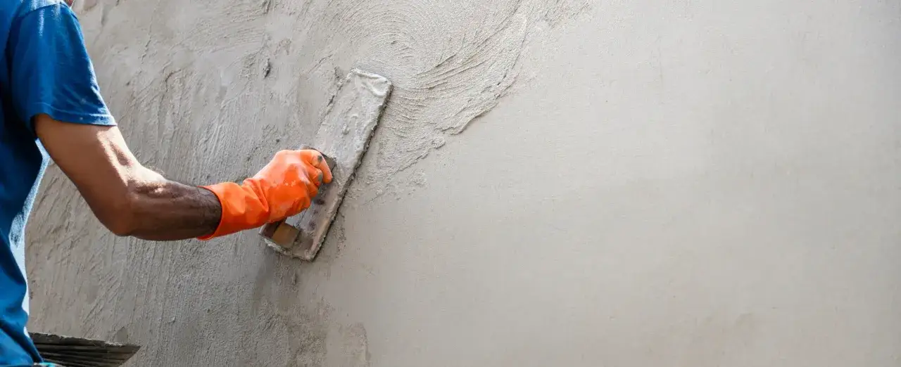 Main d'un ouvrier en gant orange appliquant de l'enduit sur agglo avec une truelle, créant une texture sur le mur.