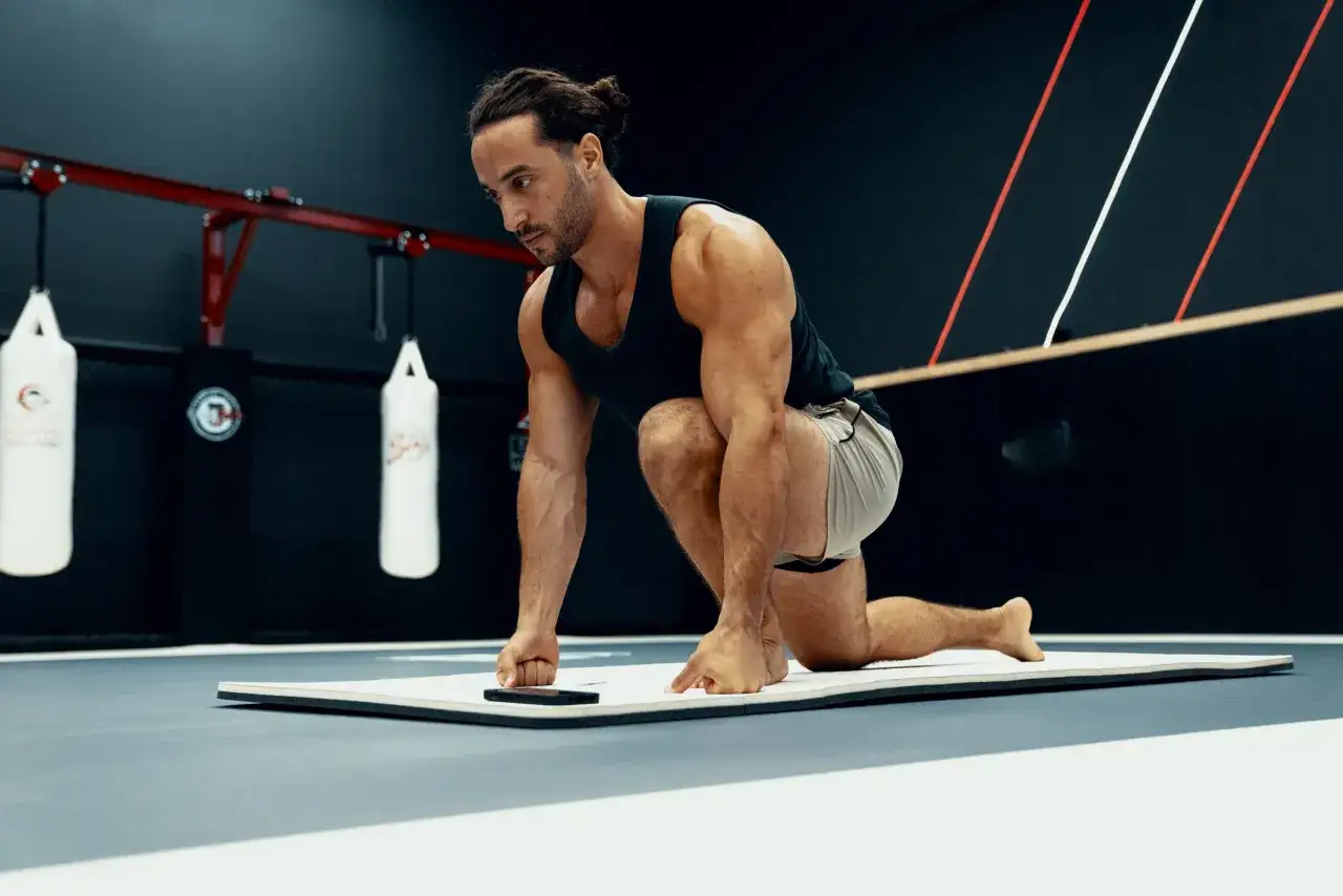 Un homme en position de fente sur un tapis, prêt pour son échauffement musculation. Des sacs de frappe sont visibles en arrière-plan.