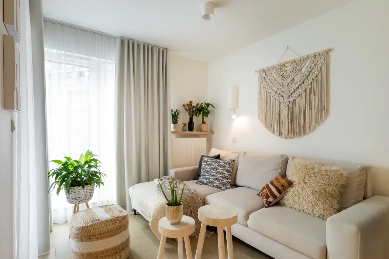 Przytulny salon w beżach i bieli. Duża, narożna kanapa, makrama na ścianie i naturalne dodatki tworzą spokojną atmosferę.
