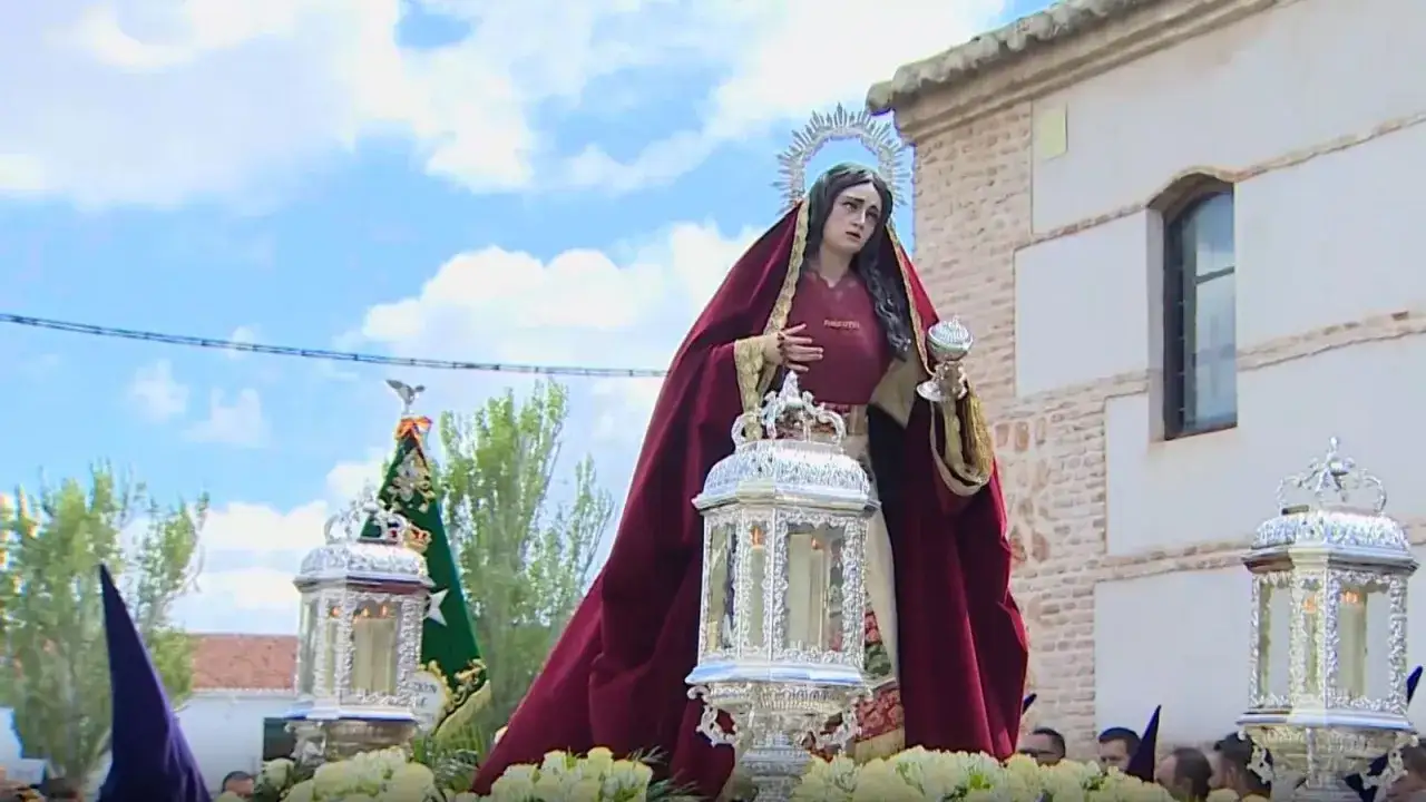 Imagen de procesiones Sábado Santo: una figura religiosa con manto rojo y halo, portando un cáliz, avanza con faroles ornamentados y flores blancas.