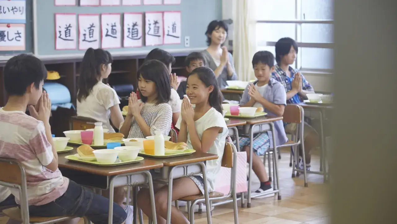 Children in a classroom practice gratitude before their Japanese school lunch. They sit at desks with trays of food, hands clasped in prayer.