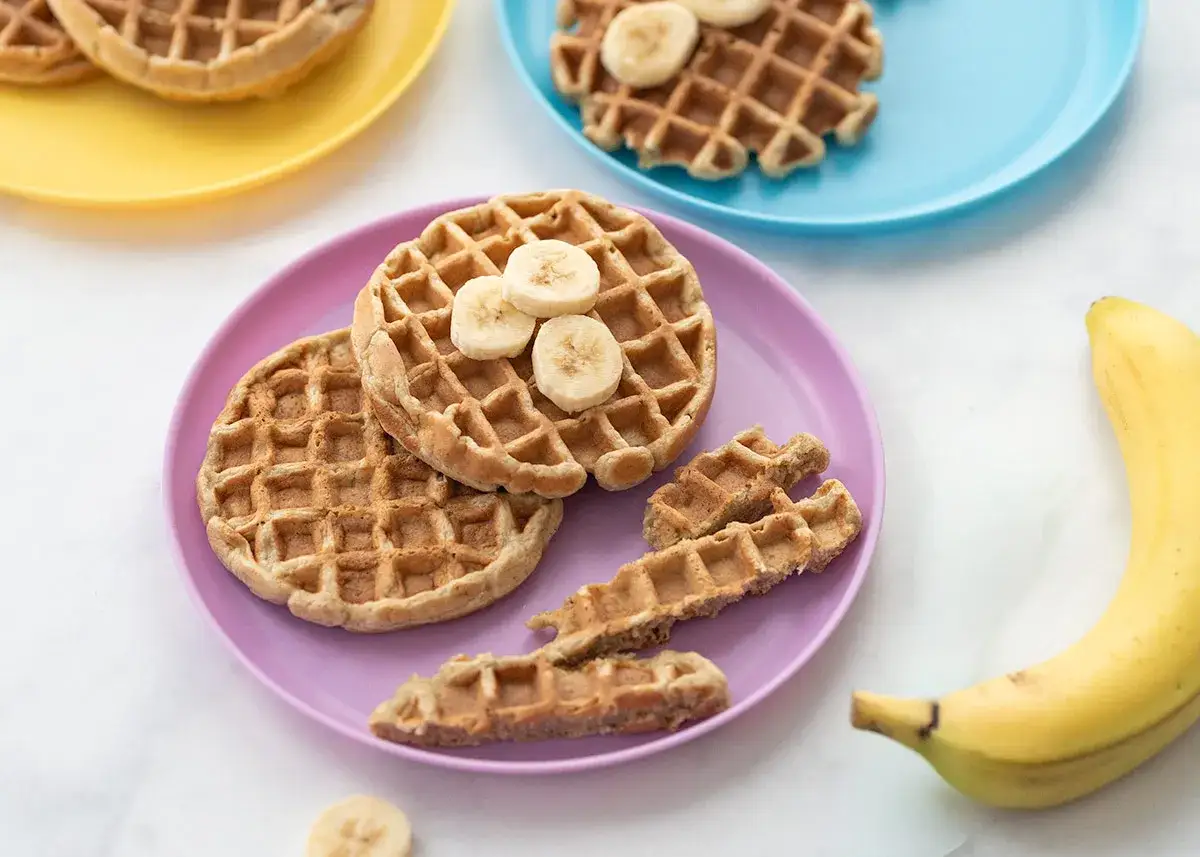 Kleine, runde Baby Waffeln mit Bananenscheiben auf einem lila Teller. Daneben liegen knusprige Waffelstäbchen und eine Banane.