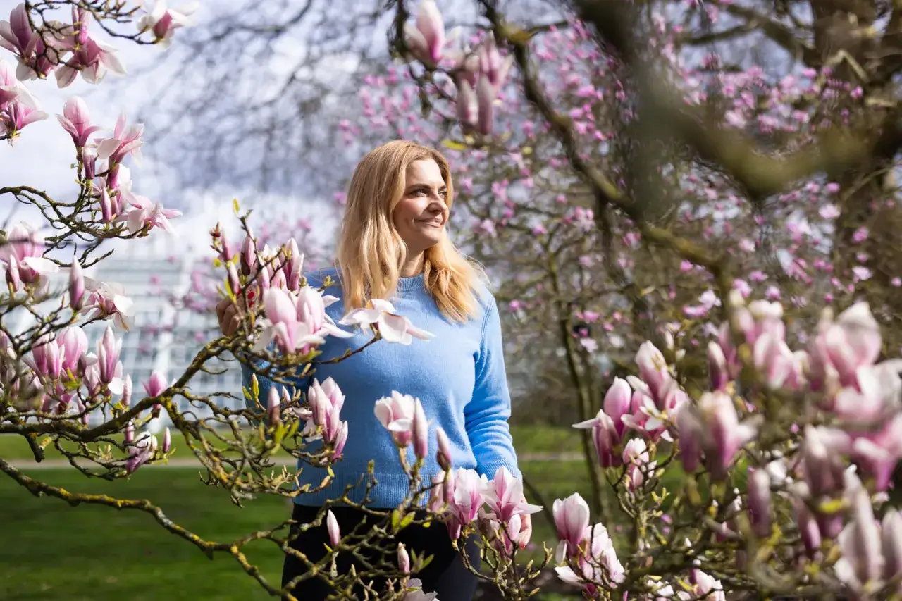 Kobieta w błękitnym swetrze wśród kwitnącej magnolii w ogrodzie. Delikatne, różowe kwiaty otaczają ją, tworząc wiosenną idyllę.