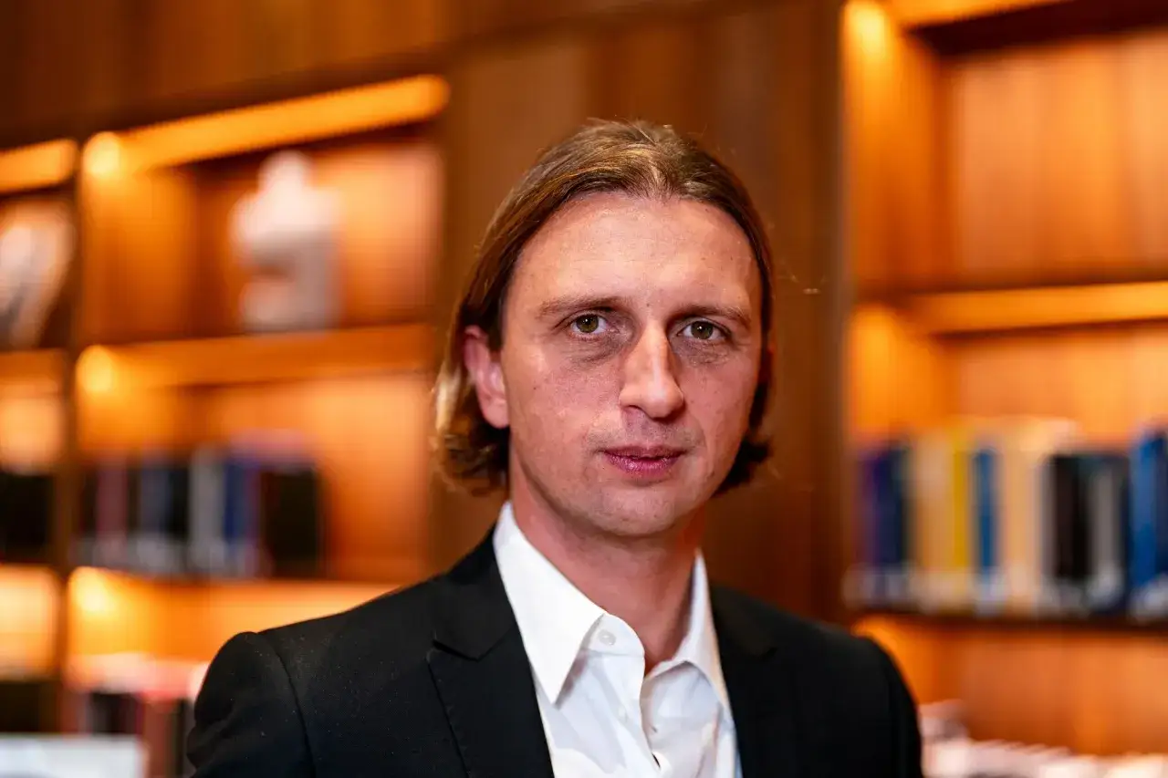 Nikołaj Storonski, zamyślony, w czarnym garniturze i białej koszuli, na tle p&oacute;łek z książkami.