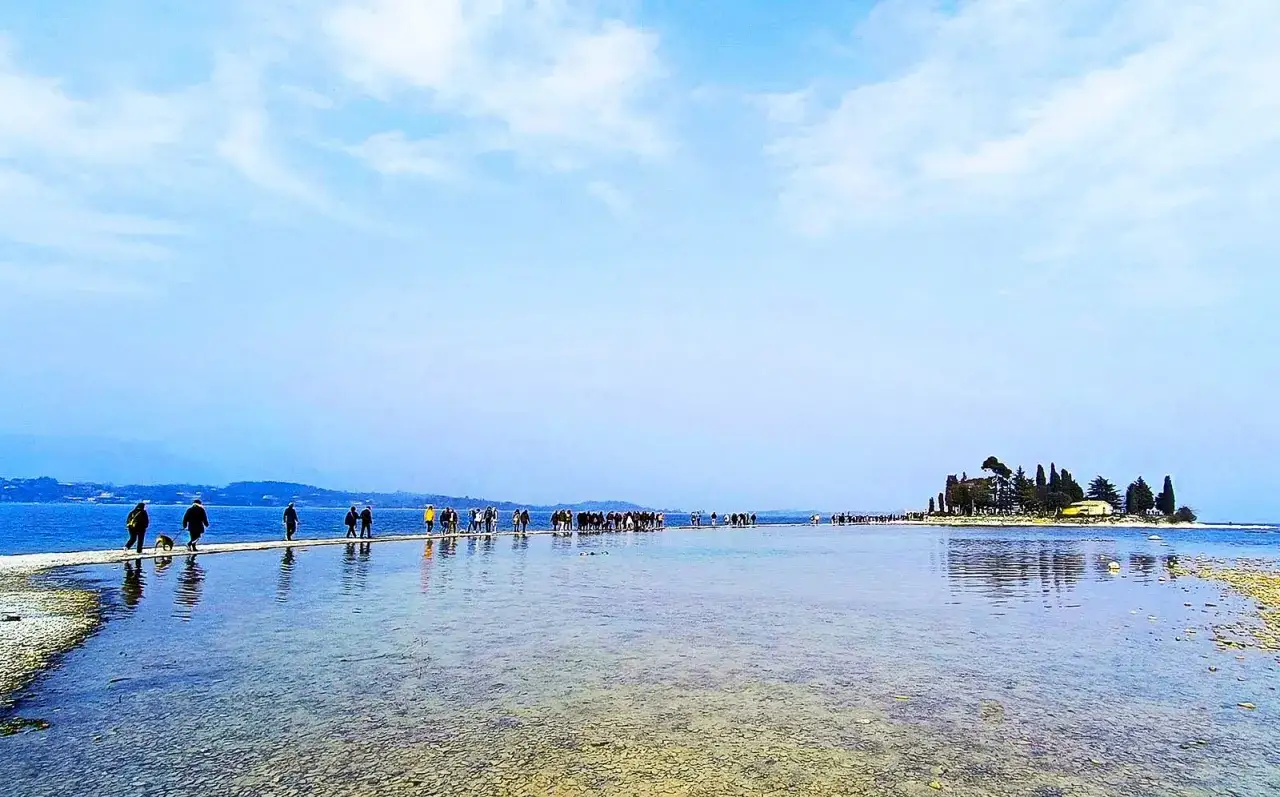 Una lunga passerella sull'acqua collega la riva a un'isola con alberi e un edificio, un'incantevole passeggiata verso l'isola dei conigli sul Lago di Garda.