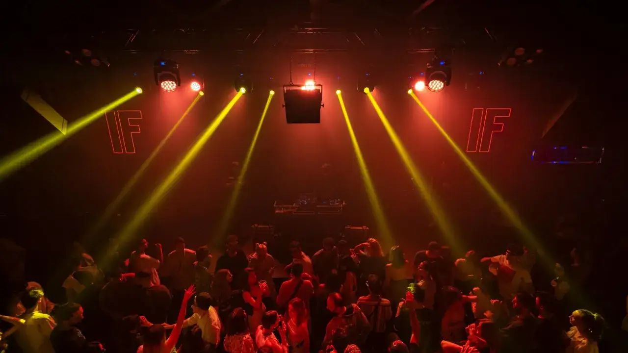 Folla in delirio in una discoteca techno, illuminata da fasci di luce gialla e rossa. Il logo 