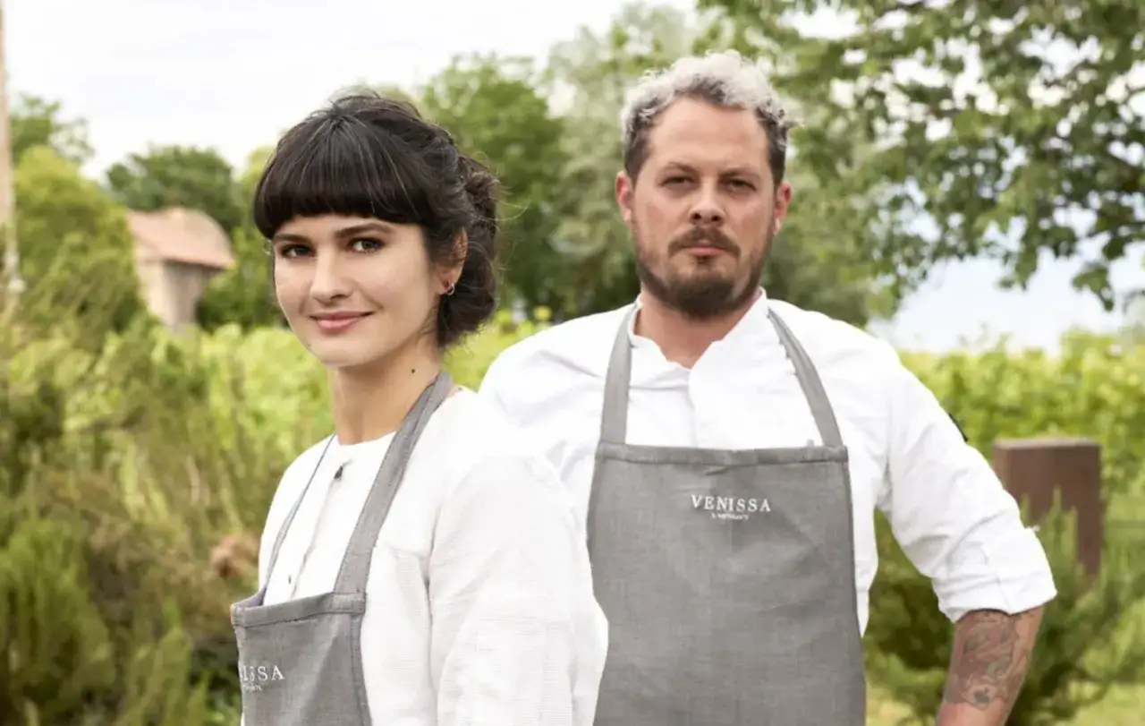 Chiara Pavan e un collega chef con grembiuli grigi, sorridono in un vigneto.