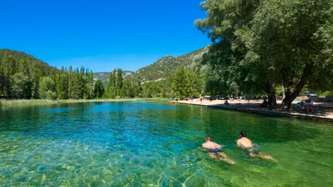 Dos hombres nadan en las aguas cristalinas de la playa de Cañamares, rodeada de vegetación y montañas.