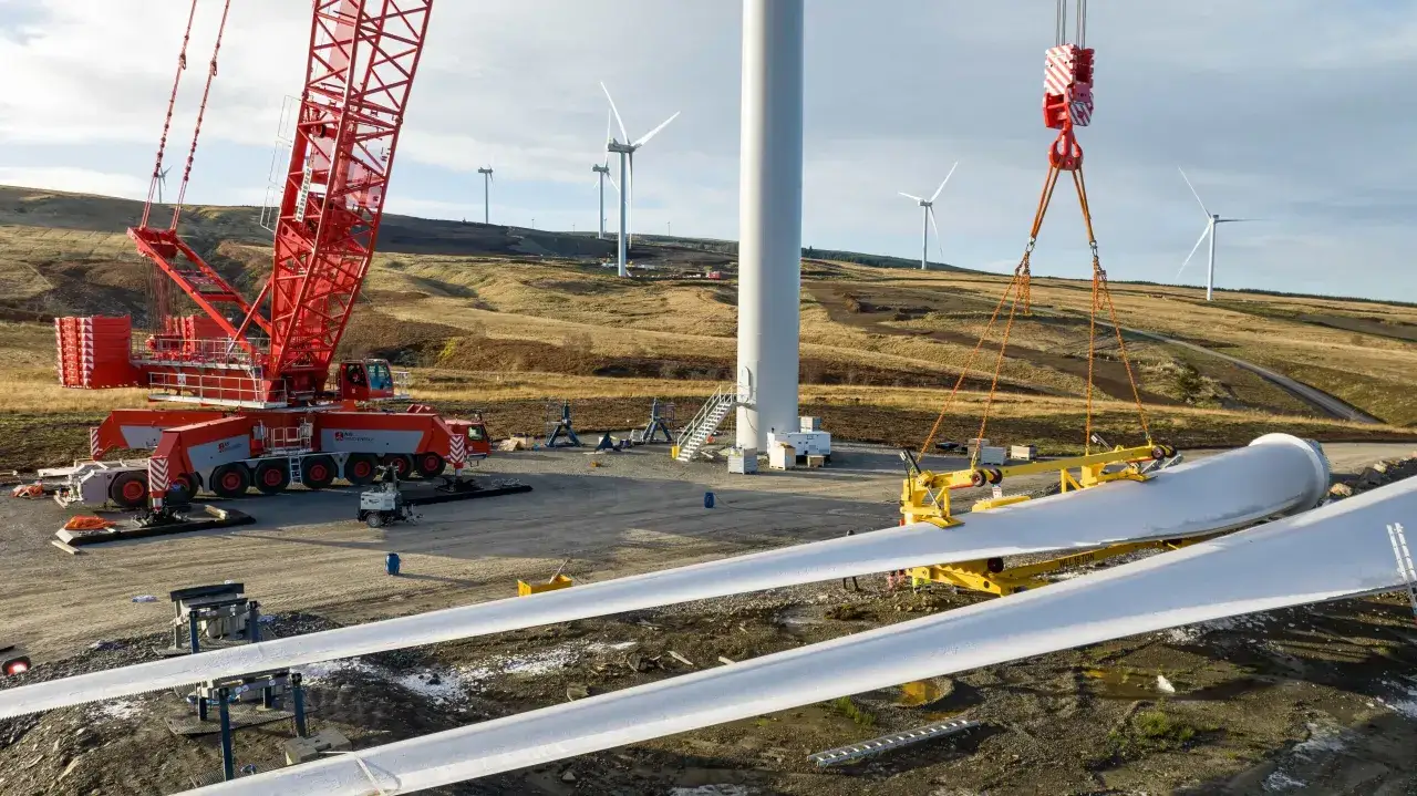 Budowa farmy wiatrowej w toku. Wielki czerwony dźwig przygotowuje się do montażu łopat wiatraka, które leżą na ziemi.